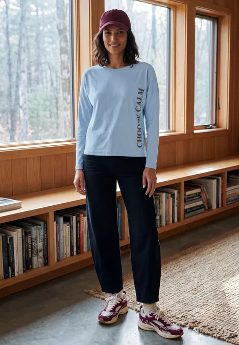 Femme en tenue décontractée (t-shirt bleu clair, pantalon foncé, baskets, casquette) devant fenêtre et bibliothèque.