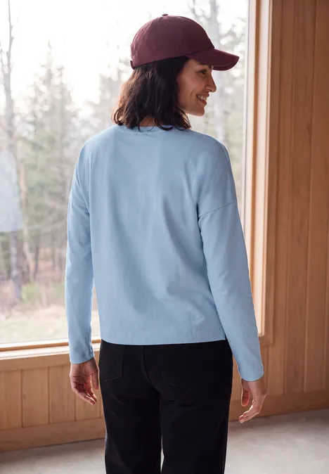Femme de dos en chemise bleu clair, pantalon foncé et casquette bordeaux, devant une grande fenêtre vers la forêt.