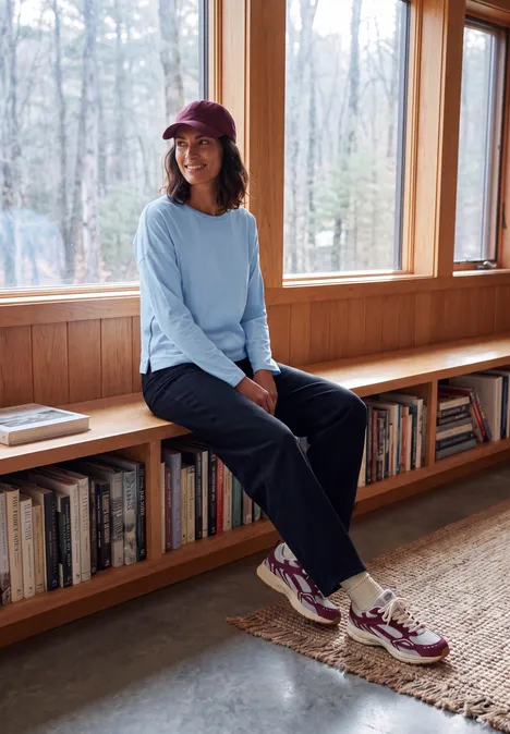 Femme en casquette et baskets assise sur un meuble en bois devant de grandes fenêtres sur la forêt.