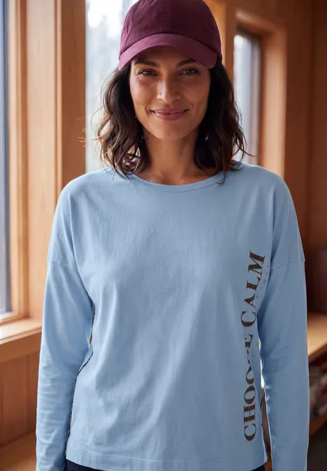 Femme souriante en t-shirt à manches longues bleu clair 'CHOOSE CALM' et casquette bordeaux près d'une fenêtre