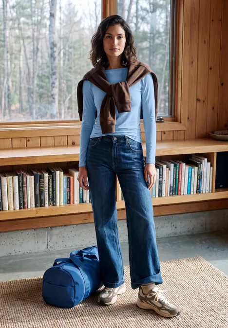 Frau in hellem Pullover, Jeans und Sneakers mit Reisetasche vor Bücherregal in Holzzimmer.