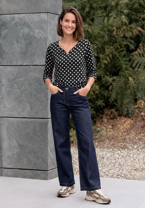 Femme souriante en chemisier noir à pois blancs, jean large bleu foncé et baskets devant un mur de pierre et des arbres.
