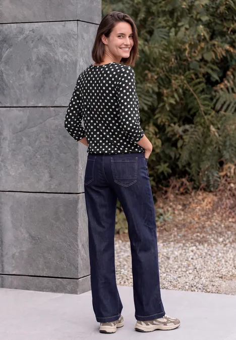 Une femme souriante en chemisier à pois et jean large se tient devant un mur gris en extérieur.
