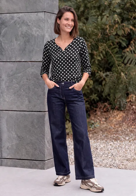 Femme en chemisier à pois, jean et baskets, souriante les mains dans les poches devant un mur gris en plein air.