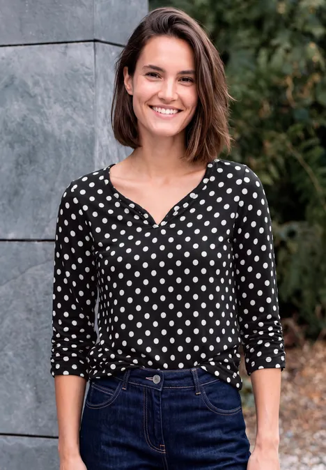 Une femme souriante en chemisier à pois et jean se tient devant un mur et des plantes.