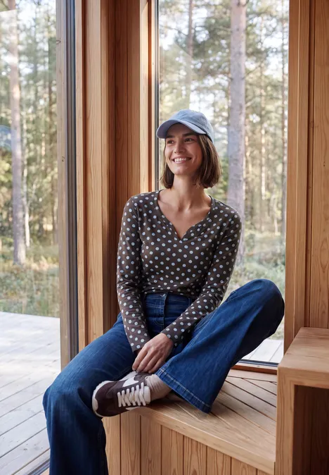 Une jeune femme en jean et chemisier à pois assise sur un banc en bois, souriante, forêt en arrière-plan.