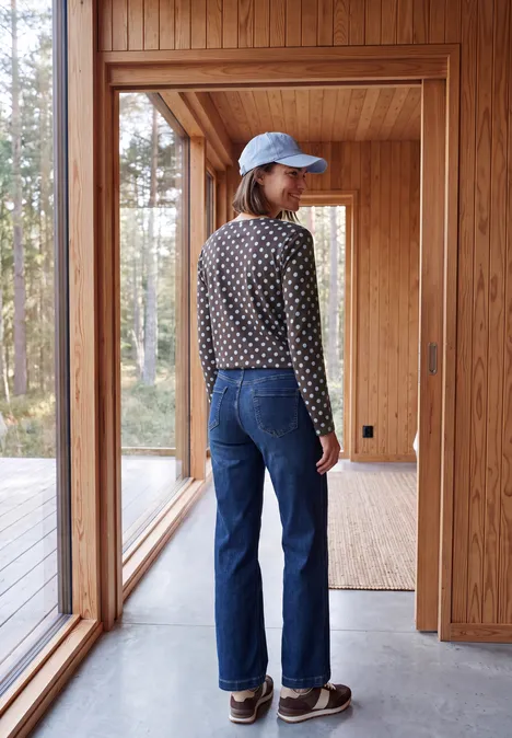 Personne portant une casquette bleue, un pull à pois et un jean se tient dans une maison en bois lumineuse, forêt visible.