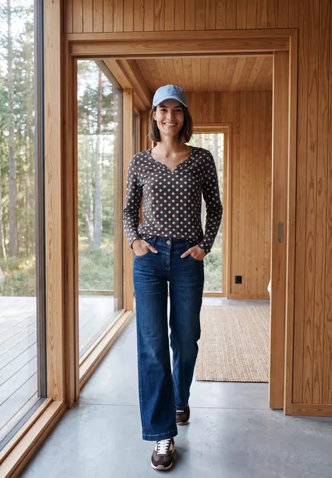 Une femme en jean, chemisier à pois et casquette se tient dans une maison moderne à bois.