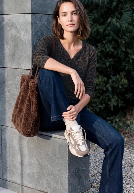 Frau sitzt lässig an einer Steinsäule, trägt gemusterten Pullover, Jeans, Sneakers und flauschige Tasche.