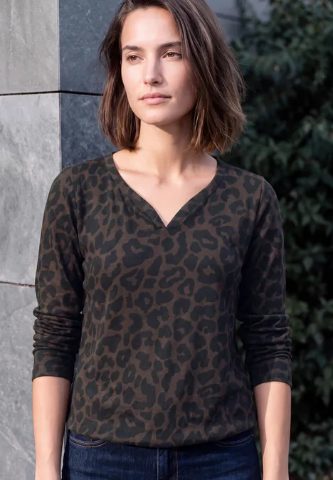 Eine Frau mit braunen Bob-Haaren trägt ein dunkelgrünes, leopardgemustertes Langarmshirt und steht vor einer Mauer.