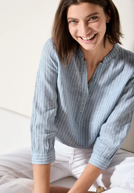 Frau lächelt, trägt blau-weiß gestreifte Bluse und weiße Hose.