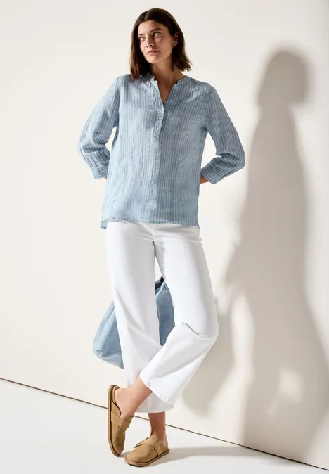 Frau trägt hellblaue Bluse, weiße Hose und beigefarbene Schuhe, steht lässig an einer Wand.