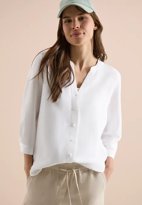 Vrouw draagt witte linnen blouse met driekwartmouwen en beige broek met koord, handen in de zakken