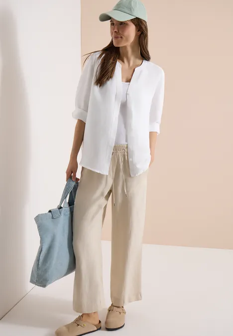 Vrouw in een zomerse, casual outfit met witte blouse, wijde beige broek, pet en blauwe tas.