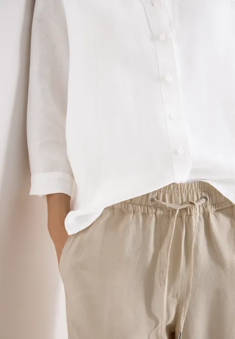 Iemand draagt een wit overhemd en een comfortabele beige broek, hand in de zak.