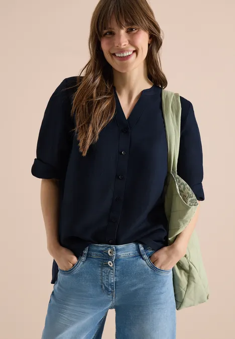 Vrouw met donkerblauw overhemd, lichtblauwe jeans en groene stoffen tas over de schouder, handen in de zakken.