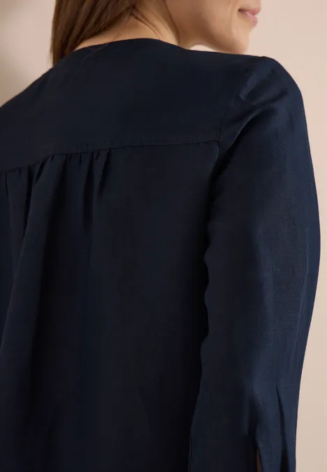 Achteraanzicht van een vrouw in een donkerblauw blouse, met lichte plooien op de rug en zichtbare schouders.