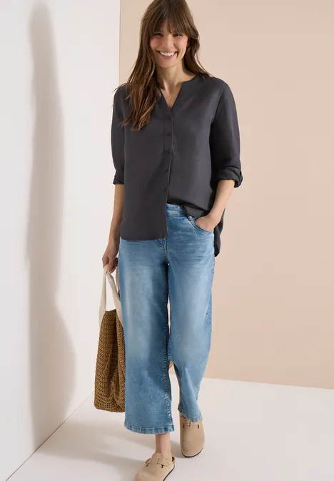 Een vrouw draagt een donkerblouse met 3/4-mouwen, wijde blauwe jeans, beige slippers en houdt een rieten tas vast.