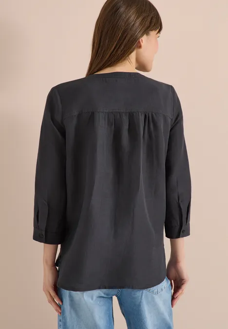 Een vrouw staat met de rug naar de camera, draagt een zwarte blouse met driekwartmouwen en een spijkerbroek.