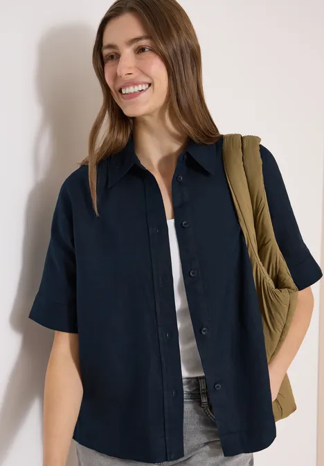 Slanke persoon draagt een donkerblauw overhemd met korte mouwen over een wit topje, jeans, olijfgroene tas.