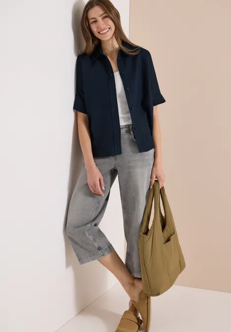 Vrouw leunt tegen lichte muur, draagt donkerblauw overhemd, wit topje, grijze 3/4 broek en kaki tas.