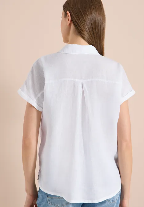 Rugaanzicht van een vrouw in een witte, korte mouwen blouse met kraag tegen een beige achtergrond.