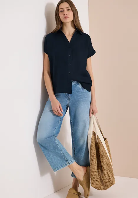 Frau lehnt an Wand, trägt schwarzes Hemd, hellblaue weite Jeans und hält einen großen geflochtenen Einkaufskorb.