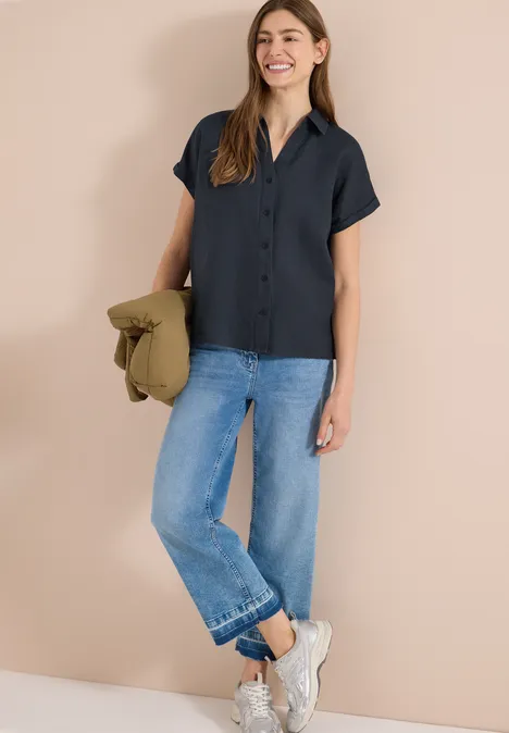 Vrouw in zwart overhemd, blauwe jeans en witte sneakers, met een kaki jasje.