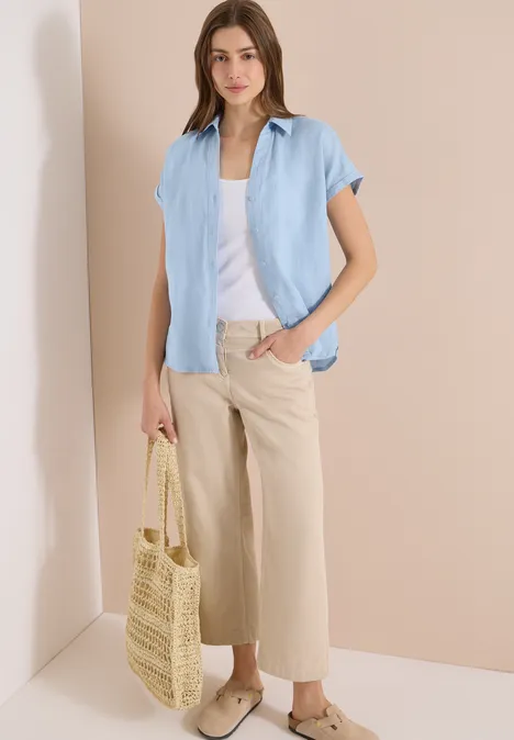 Vrouw in lichtblauw overhemd, wit topje, beige wijde broek en strooien tas.