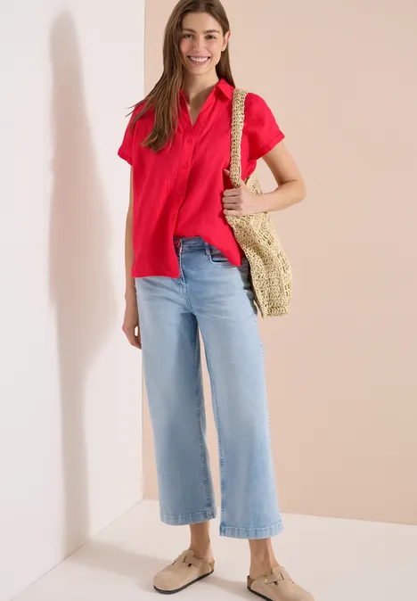 Vrouw met rood poloshirt, lichtblauwe wijdvallende jeans, rieten tas en beige espadrilles tegen lichte achtergrond.
