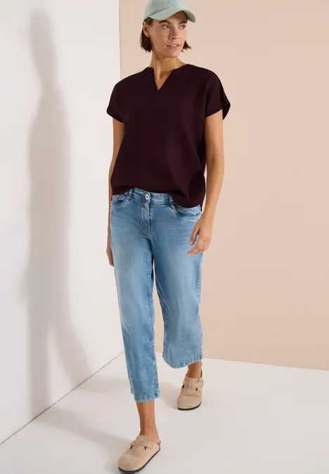 Frau trägt dunkelviolettes V-Neck-T-Shirt, weite 3/4-Jeans und beige Sneakers vor neutralem Hintergrund.
