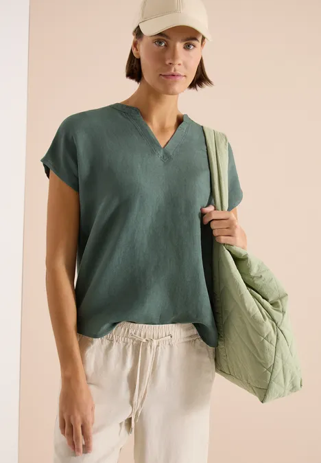 Person in lockerem, grünem V-Shirt und beigen Leinenhosen mit großer, gesteppter, grüner Umhängetasche.