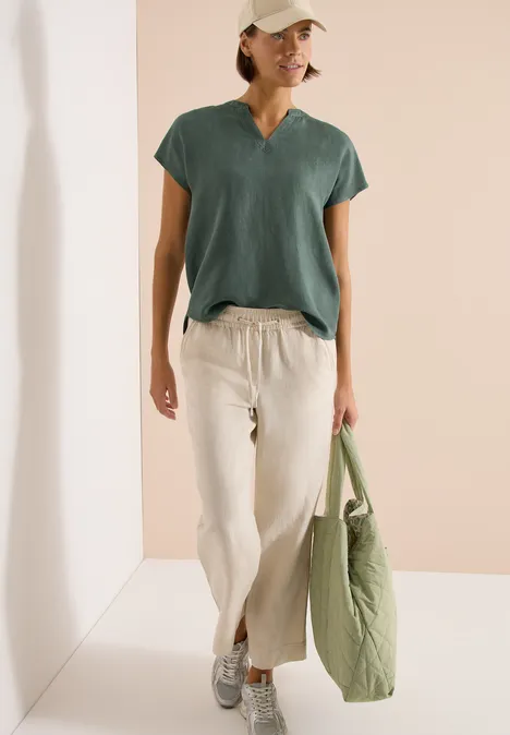 Person vor hellem Hintergrund in lockerem Outfit: grünes Shirt, helle Leinenhose, Sneakers und grüne Tasche.