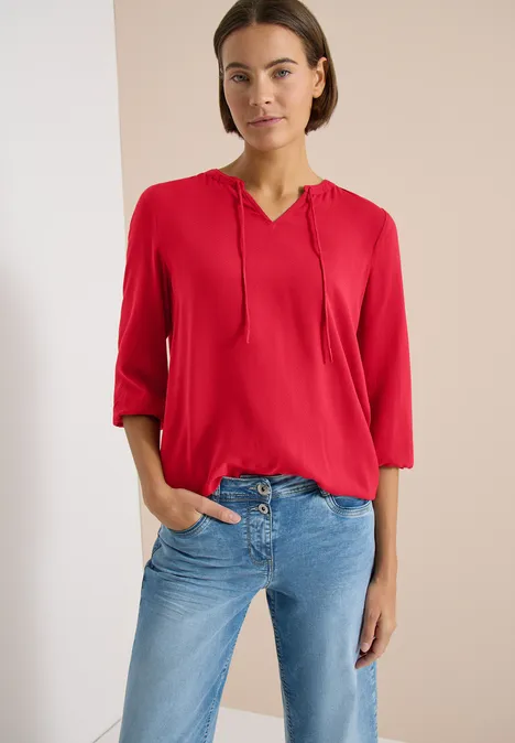 Femme en chemise rouge à capuche et jean bleu clair, posant devant un fond neutre.