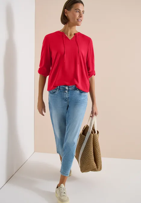 Femme en chemisier rouge et jean bleu clair appuyée contre un mur, avec un sac tressé.