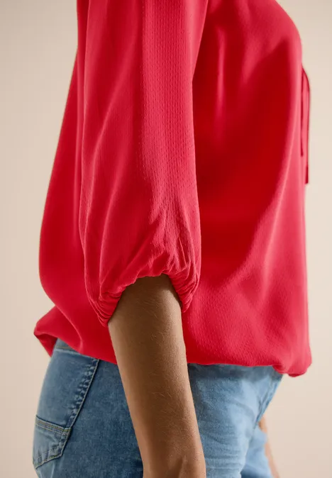 Vue de profil d'une personne portant un chemisier rouge à manches 3/4 et un jean bleu.