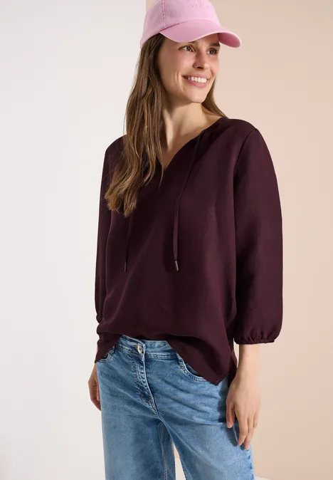 Vrouw in bordeaux hoodie, spijkerbroek en roze pet voor lichte achtergrond.