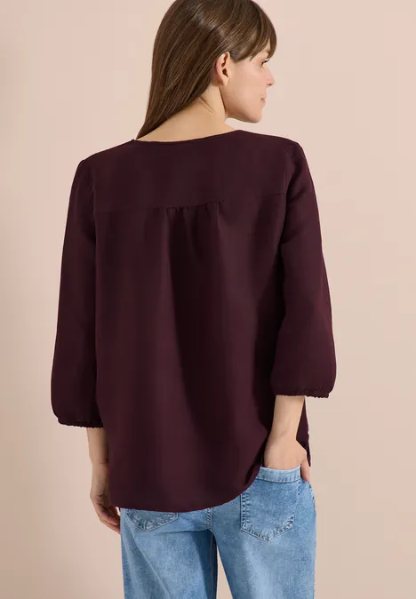 Vrouw gezien van achteren in een bordeaux shirt met driekwarts mouwen en een spijkerbroek, beige achtergrond.