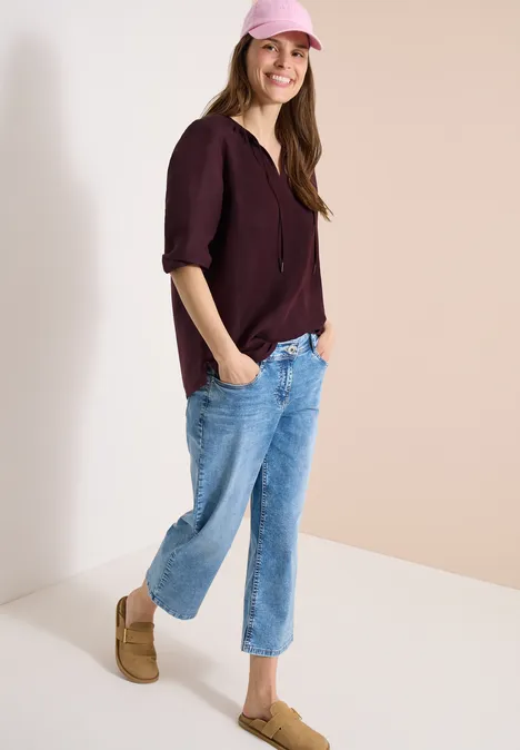 Vrouw in bordeaux top, blauwe 7/8-jeans en beige mocassins, ontspannen tegen een hoek leunend.