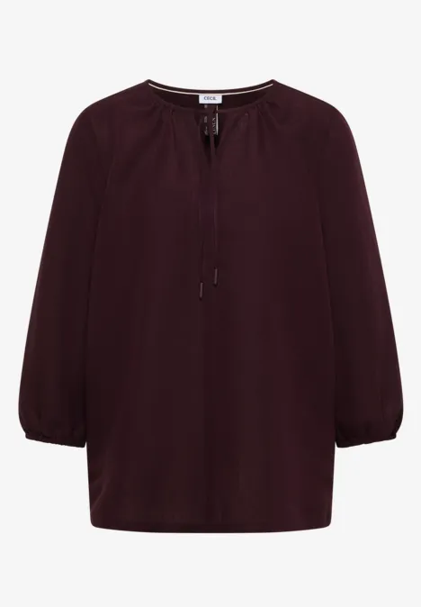 Bordeauxrode blouse met driekwartsmouwen en strikdetail bij de hals.