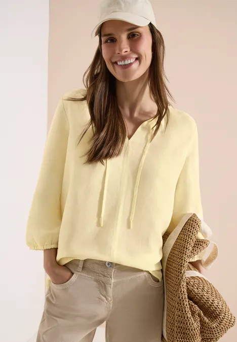 Vrouw in lichtgele blouse met striksluiting en beige broek, houdt gevlochten tas vast.