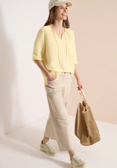 Vrouw in gele blouse, beige wijde broek, sneakers en strozak voor een neutrale achtergrond.