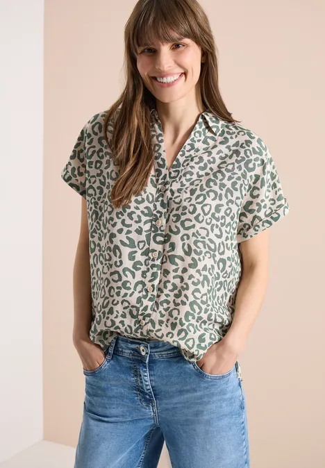 Vrouw in kort mouw luipaardprintblouse en spijkerbroek met handen in zakken, tegen effen achtergrond.