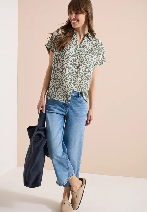 Vrouw in groen dessin overhemd met korte mouwen, blauwe jeans, beige loafers en een donkerblauwe tas.