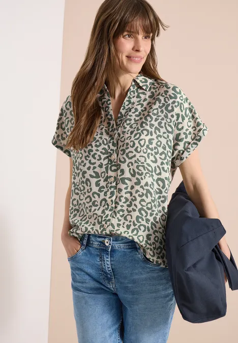 Vrouw met groen leopardshirt, jeans en een jas in de hand voor een lichte achtergrond.