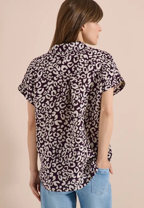 Een vrouw van achteren draagt een blouse met zwart-witte luipaardprint en lichtblauwe jeans voor een beige achtergrond.