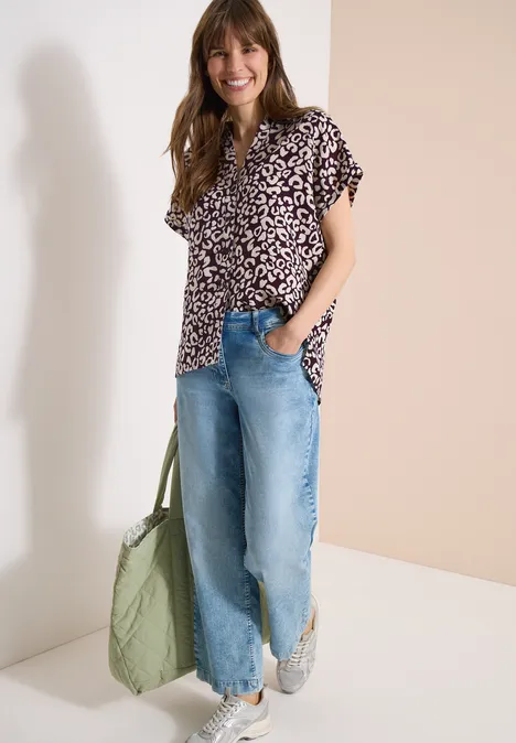 Een vrouw in een bloemenblouse, wijde spijkerbroek, witte sneakers en een groene gewatteerde tas.