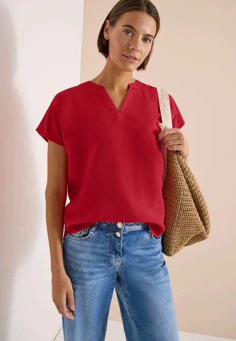 Persoon draagt rood shirt met korte mouwen en blauwe jeans, met gevlochten tas over schouder voor neutrale achtergrond.