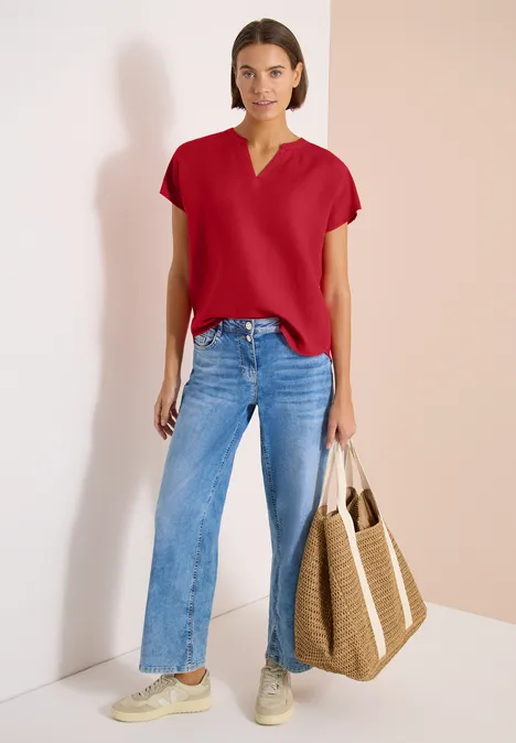 Vrouw voor lichte achtergrond met rood shirt, blauwe jeans, sneakers en een gevlochten strooien tas.