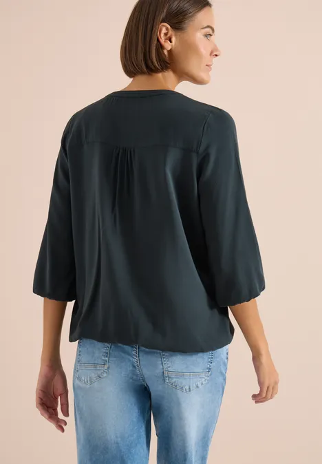 Achteraanzicht van een vrouw in een losse donkere blouse en lichtblauwe jeans, tegen een beige achtergrond.